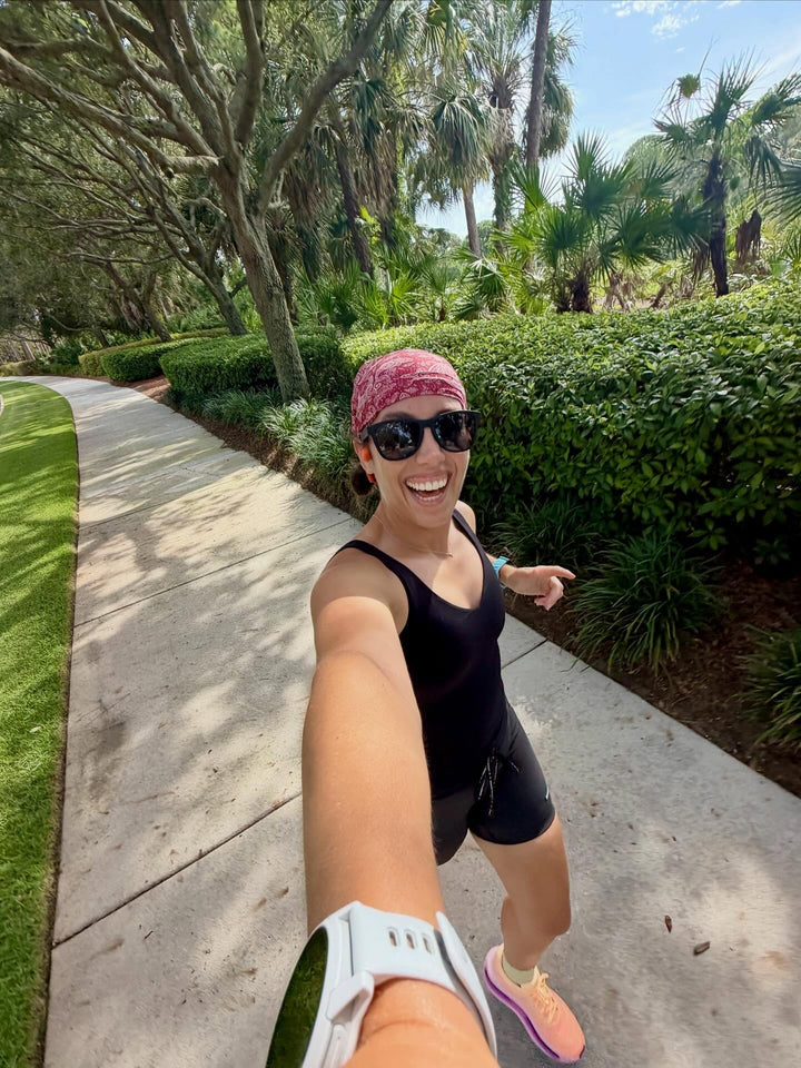 A woman wearing a pink patterned headband, black tank top, black shorts, and pink running shoes takes a selfie on a paved path surrounded by green hedges and palm trees.