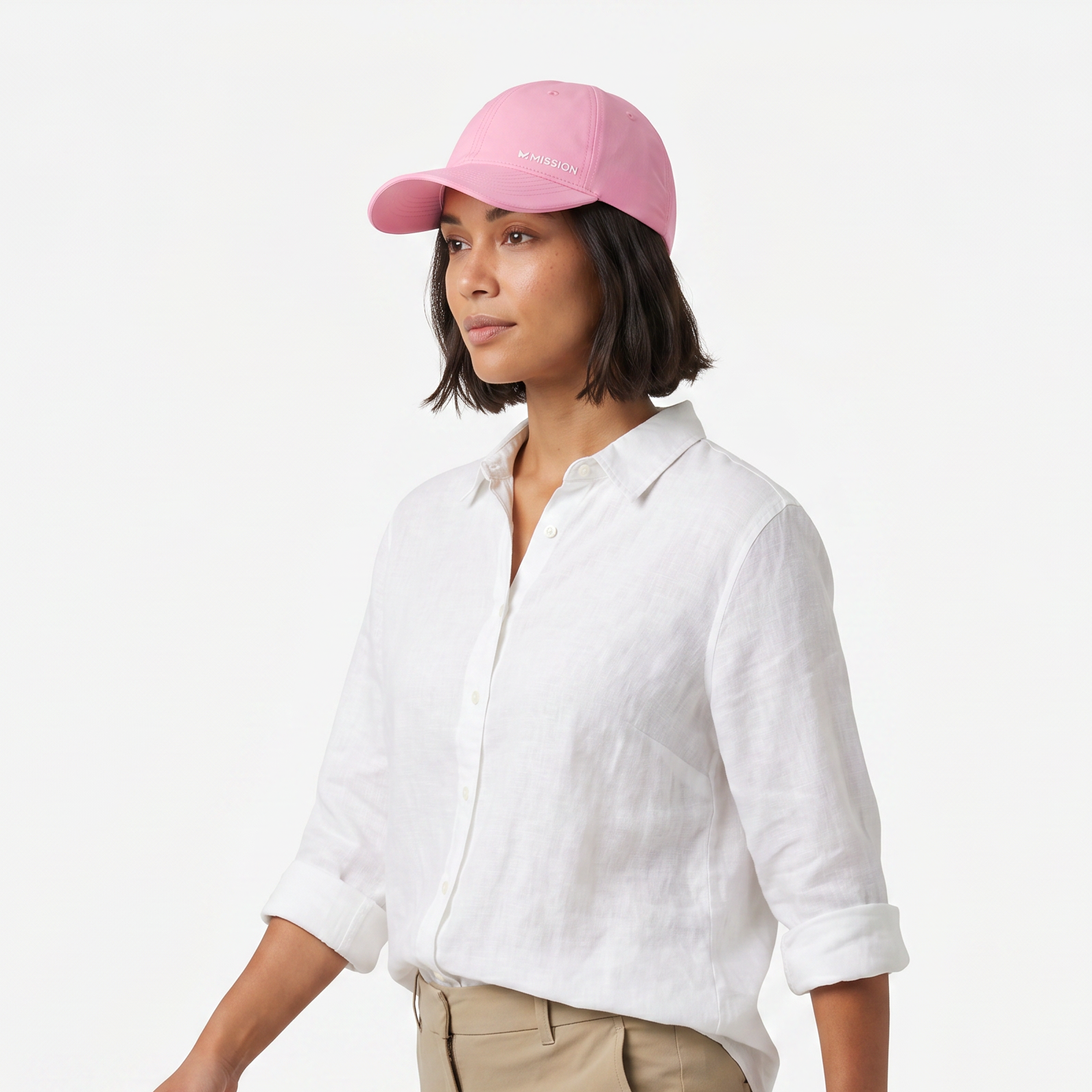 A woman wearing a light pink baseball cap with white embroidery, a white linen button-down shirt, and beige trousers, photographed against a clean white studio background.