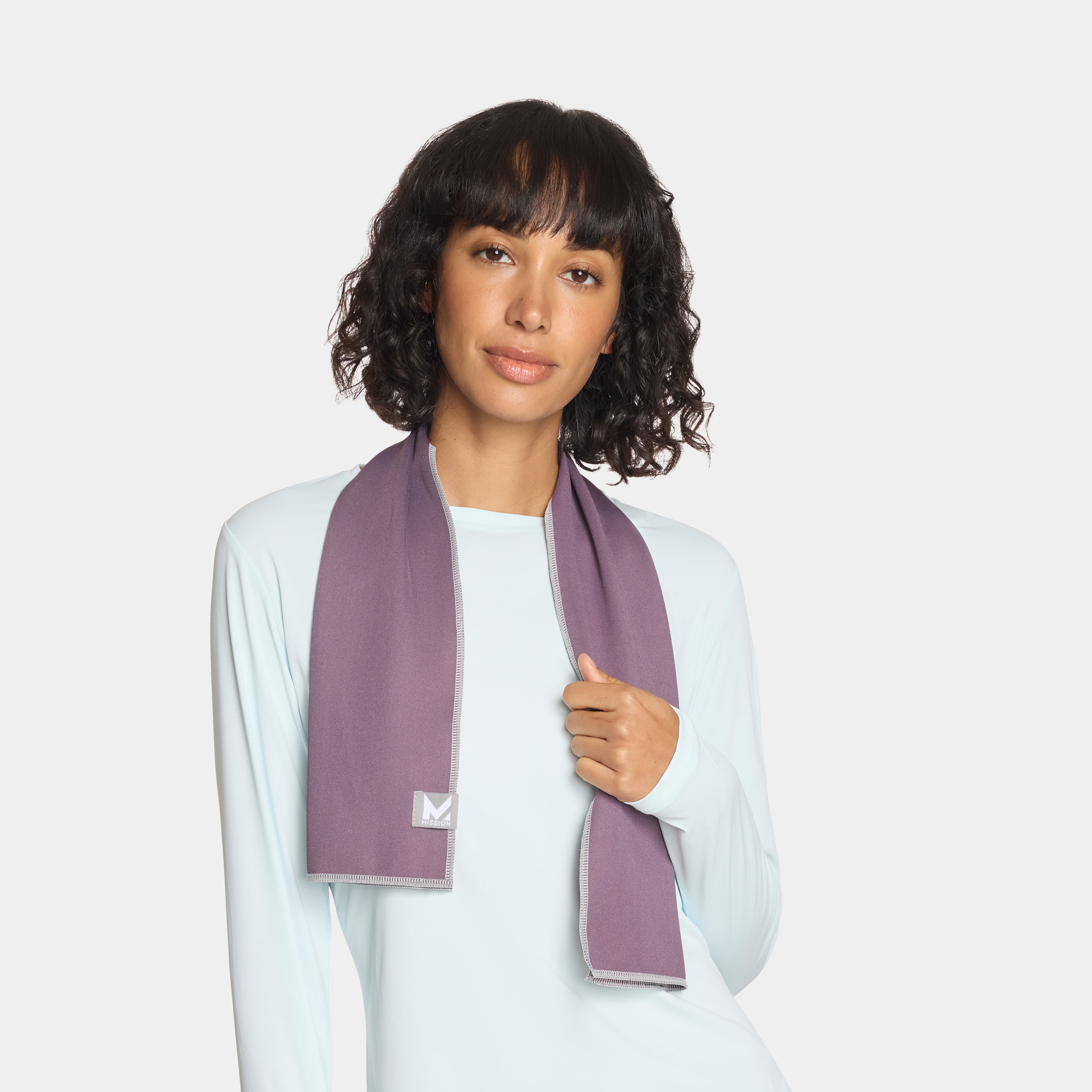 A woman with dark curly hair wears a light blue long-sleeved shirt and a purple textured scarf draped around her neck against a plain white background.