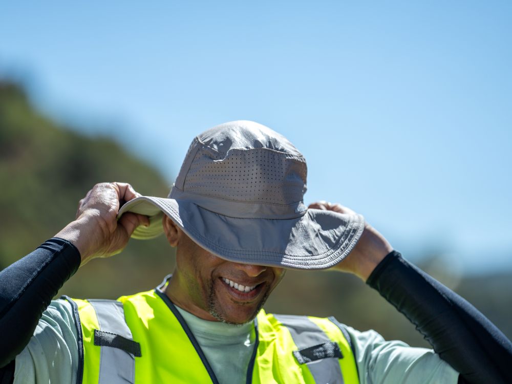A man outdoors wearing a light gray wide-brim sun hat and a high-visibility yellow safety vest, smiling as he adjusts the brim of the hat.