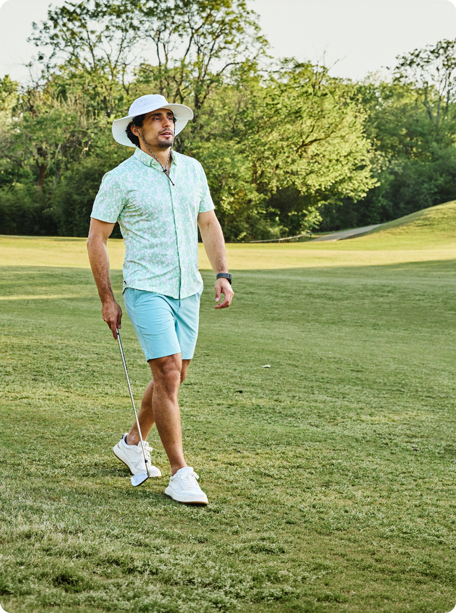 A man walking on a golf course wearing a light green and white patterned short-sleeve button-up shirt, light blue shorts, a white sun hat, and white golf shoes, holding a golf club.