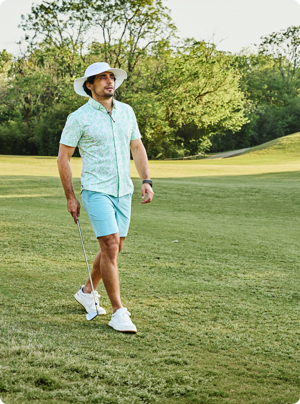 A man walking on a golf course wearing a light green and white patterned short-sleeve button-up shirt, light blue shorts, a white sun hat, and white golf shoes, holding a golf club.