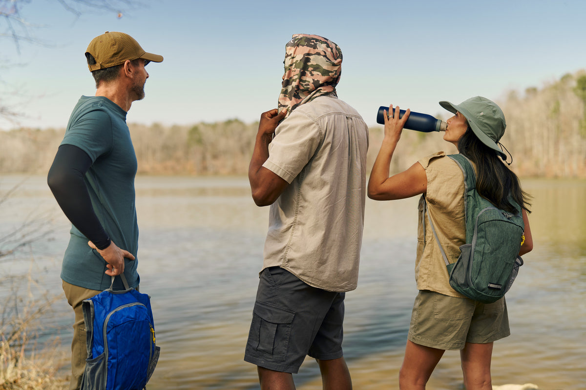 Three outdoor adventurers standing by a lake, with one person drinking from a blue water bottle, another wearing a camouflage neck gaiter, and a third carrying a blue backpack.