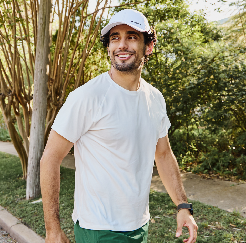 Man in a white t-shirt, white cap, and smartwatch smiles while walking outdoors on a sunny day, with trees and greenery in the background.