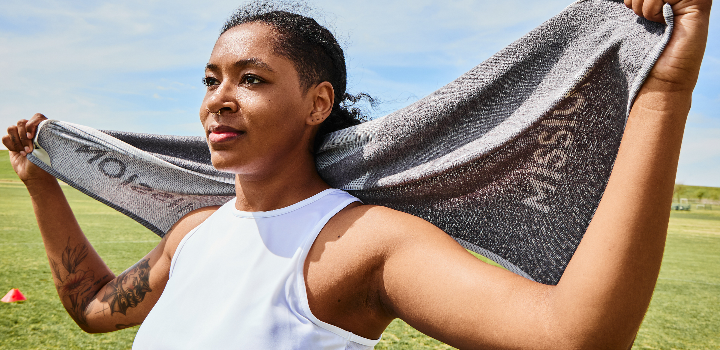 Athletic Black woman in a white sleeveless top holds a gray towel behind her on a sunny sports field, conveying confidence and strength.