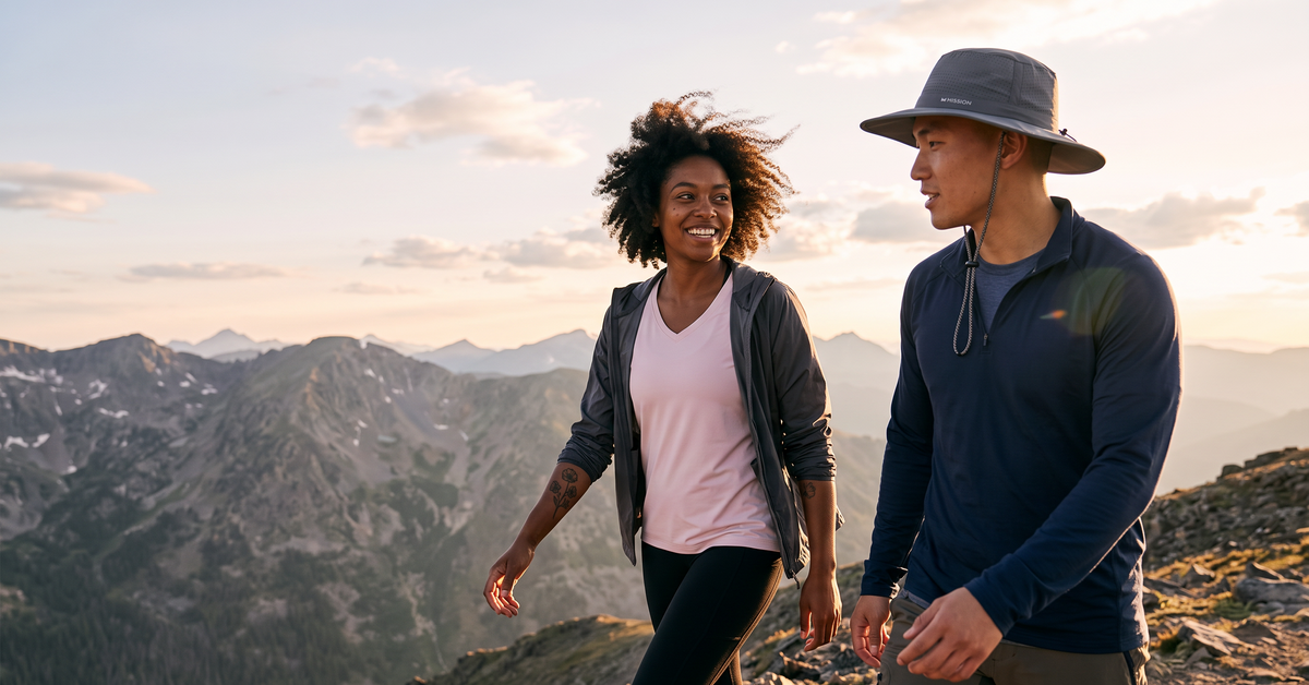 Two people hiking on a mountain ridge during golden hour, with rugged peaks and clouds in the background. One wears a wide-brim gray hat, navy quarter-zip fleece, and olive pants, while the other wears a light pink top, dark gray zip jacket, and black leggings.