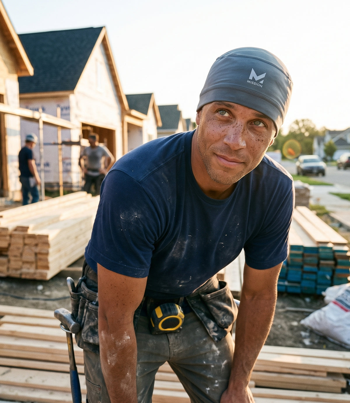A male construction worker wearing a dark blue t-shirt, gray work pants, and a gray beanie with a white 'M-Logic' logo, looking directly at the camera on a construction site at sunset.